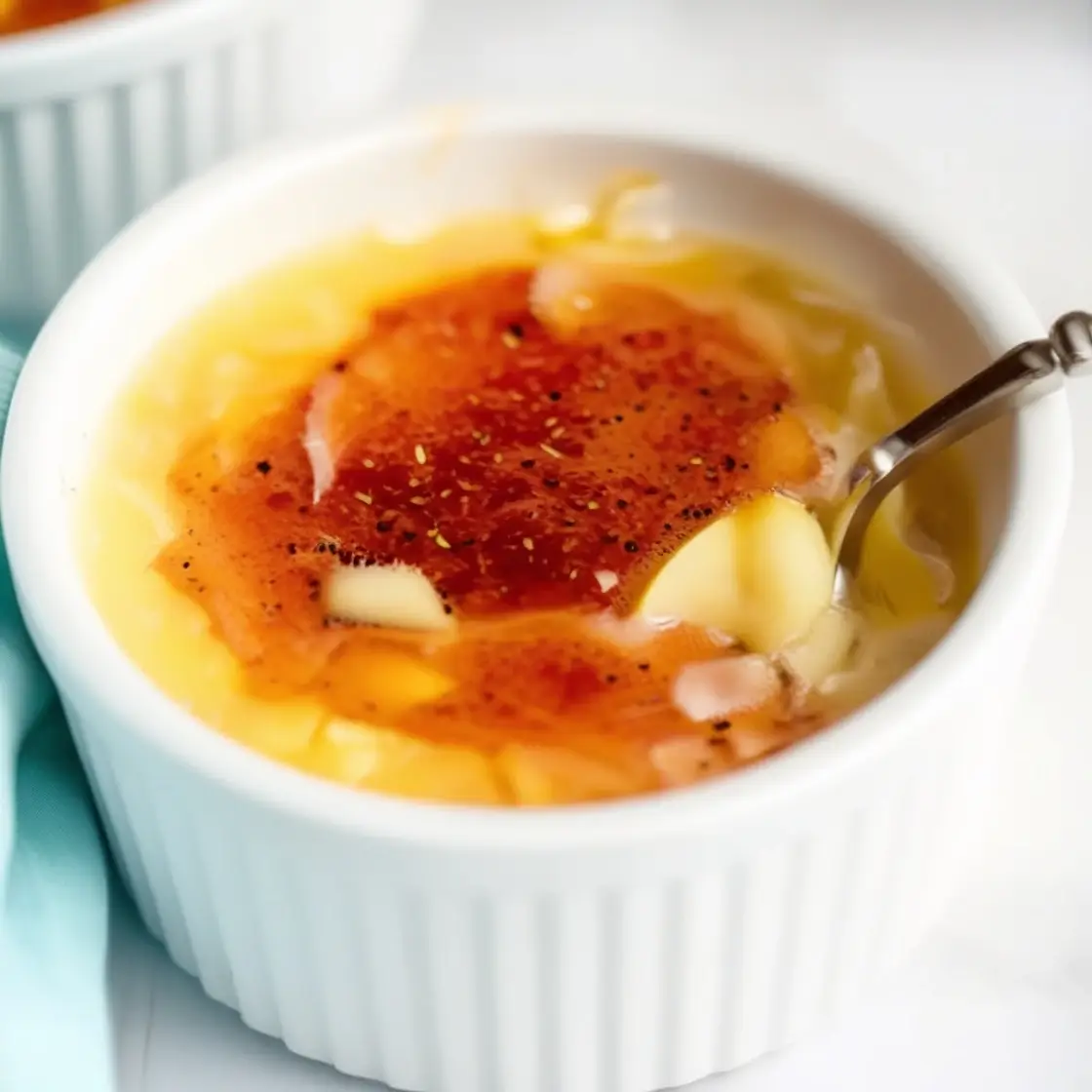 A close-up view of a creamy crème brûlée in a white ramekin, featuring a golden caramelized sugar topping cracked open with a spoon.