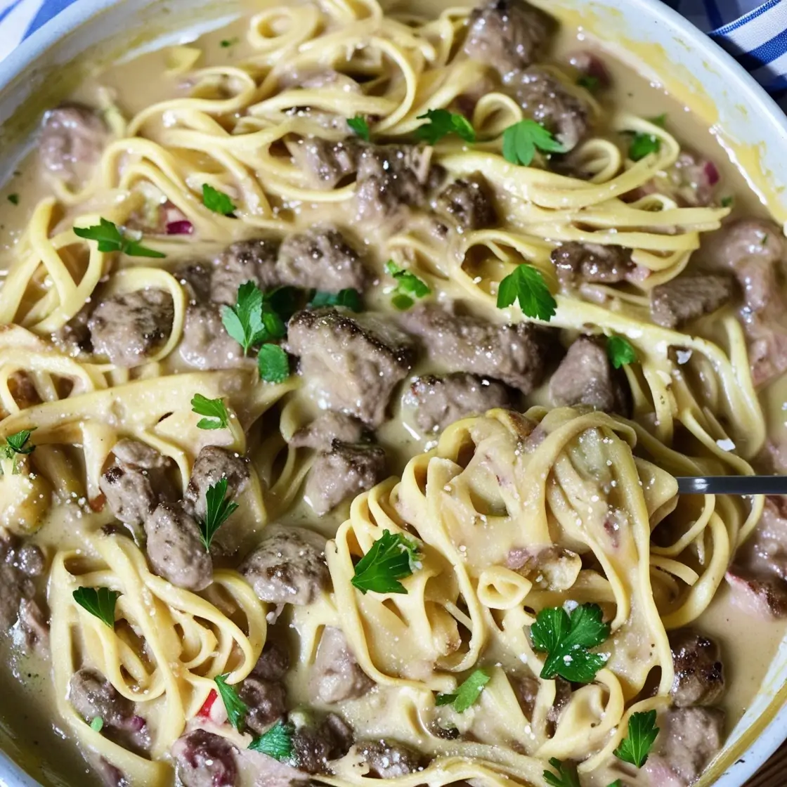 A creamy plate of pasta with ground beef in Alfredo sauce, topped with fresh parsley and parmesan, served on a striped tablecloth.